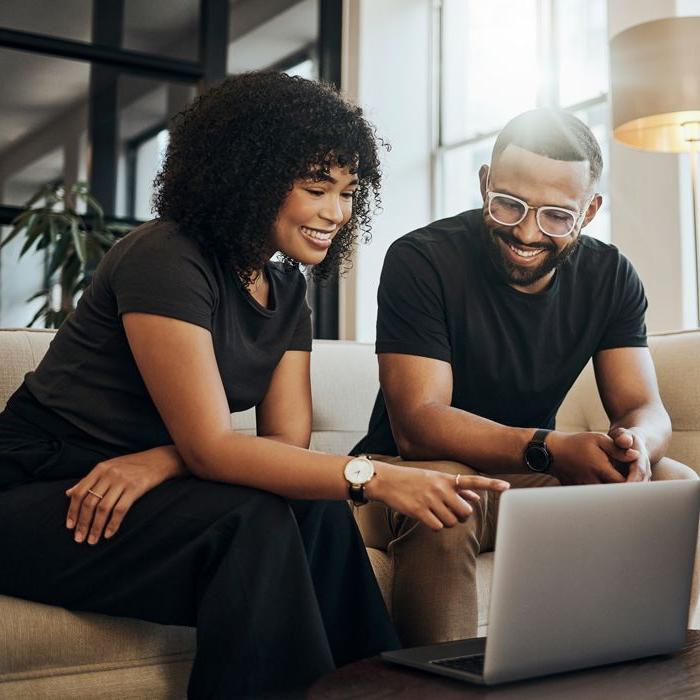 Two Patelco members sitting on a couch and researching options for local resources, both pointing at a laptop and smiling